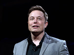 Elon Musk in a Grey Suit and Black Shirt Speaking on Stage Against a Dark Background HD wallpaper