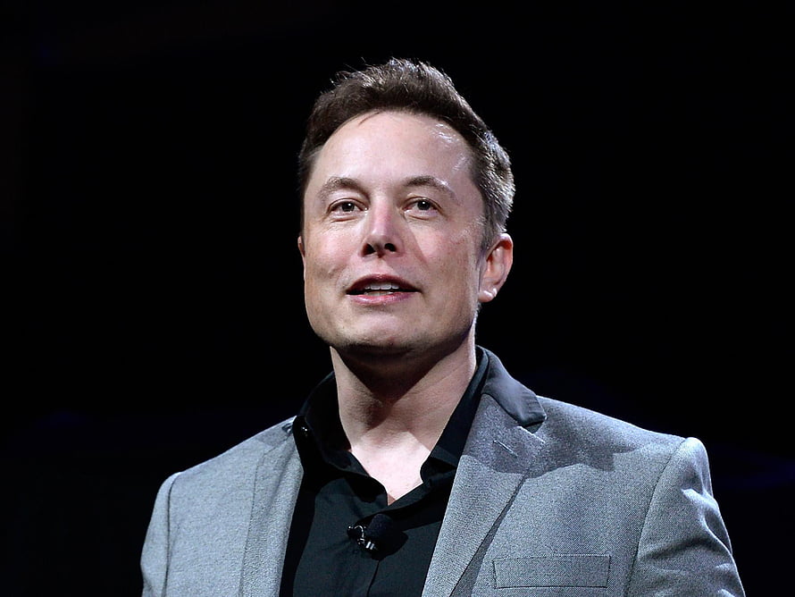 Elon Musk in a Grey Suit and Black Shirt Speaking on Stage Against a Dark Background HD wallpaper