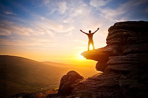 Silhouette of a person standing on a rocky cliff edge with arms raised against a golden sunset sky HD wallpaper