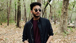 Elvish Yadav in a black bomber jacket and sunglasses standing in a forest HD wallpaper