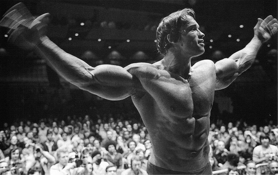 Arnold Schwarzenegger posing with arms raised at a bodybuilding competition in black and white HD wallpaper
