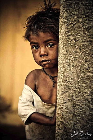 Portrait of a young child with expressive eyes peeking from behind a textured concrete pillar HD phone wallpaper