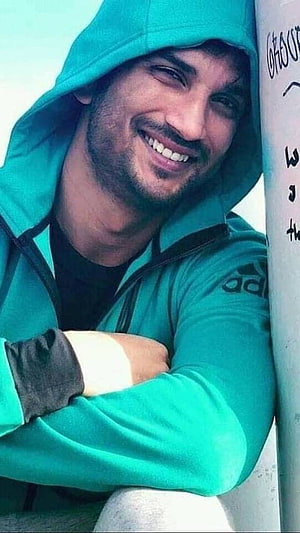 Sushant Singh Rajput smiling in a teal hooded jacket leaning against a pole HD phone wallpaper