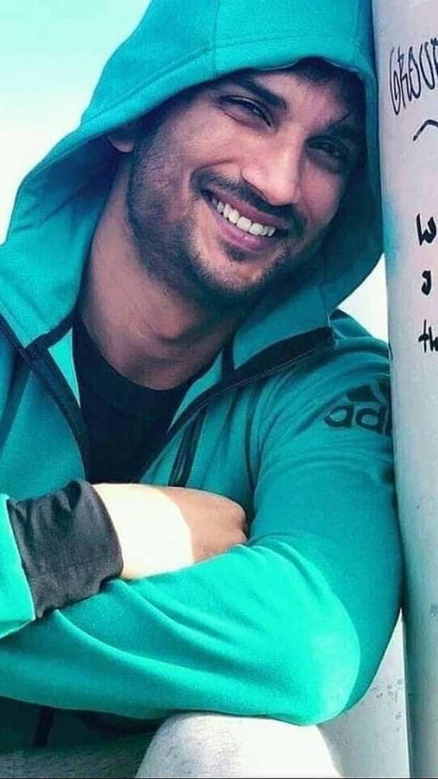 Sushant Singh Rajput smiling in a teal hooded jacket leaning against a pole HD phone wallpaper
