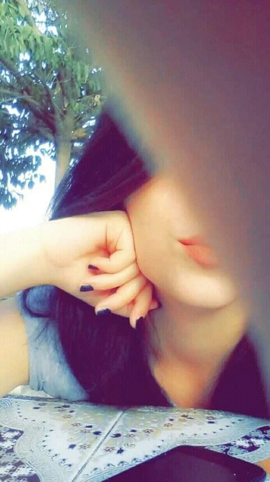Close-up portrait of a girl with dark hair resting her chin on her hand, featuring black nail polish and a blurred face, set against a background of green trees and a patterned tablecloth. HD phone wallpaper