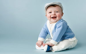 Smiling baby boy in blue cardigan and cap sitting on pastel blue studio background HD wallpaper