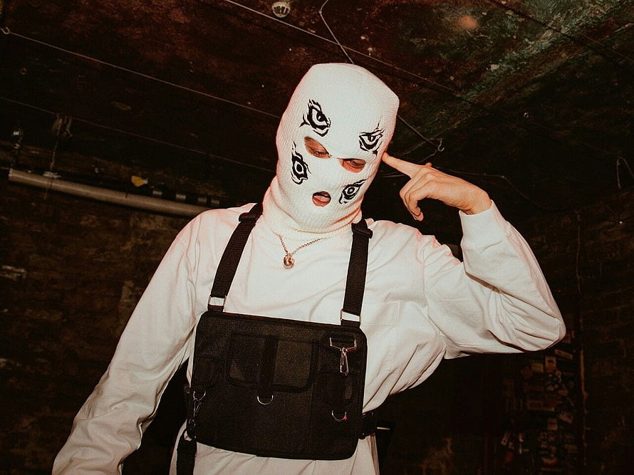 Person in white ski mask with eye designs and black chest rig in dark industrial setting HD wallpaper