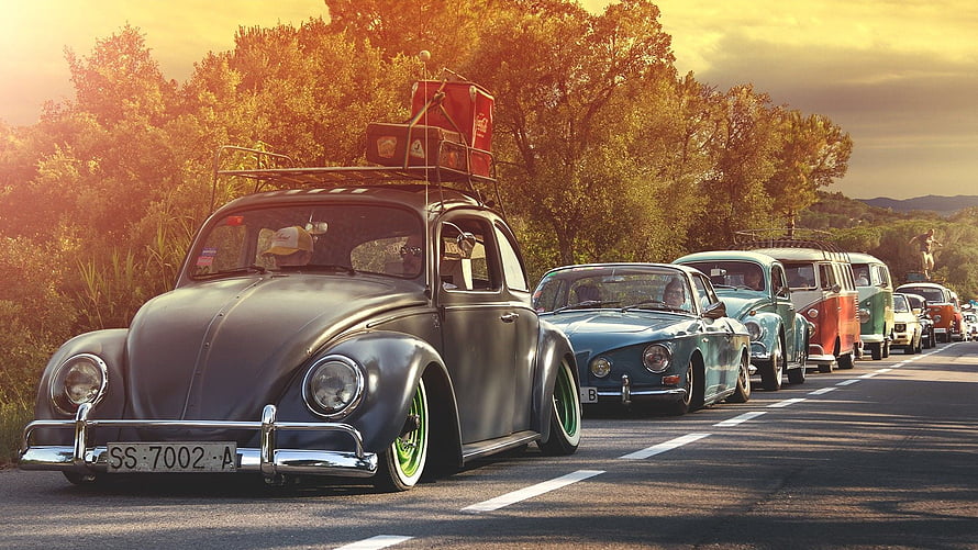 Classic Volkswagen Beetle convoy on a scenic road at golden hour with vintage cars and warm sunset light HD wallpaper