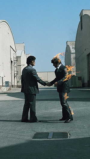 Two men in suits shaking hands in an industrial courtyard while one is engulfed in flames HD phone wallpaper