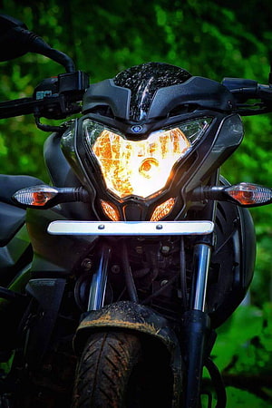 Close-up of a black NS200 motorcycle front headlight with warm amber glow against a lush green forest backdrop HD phone wallpaper