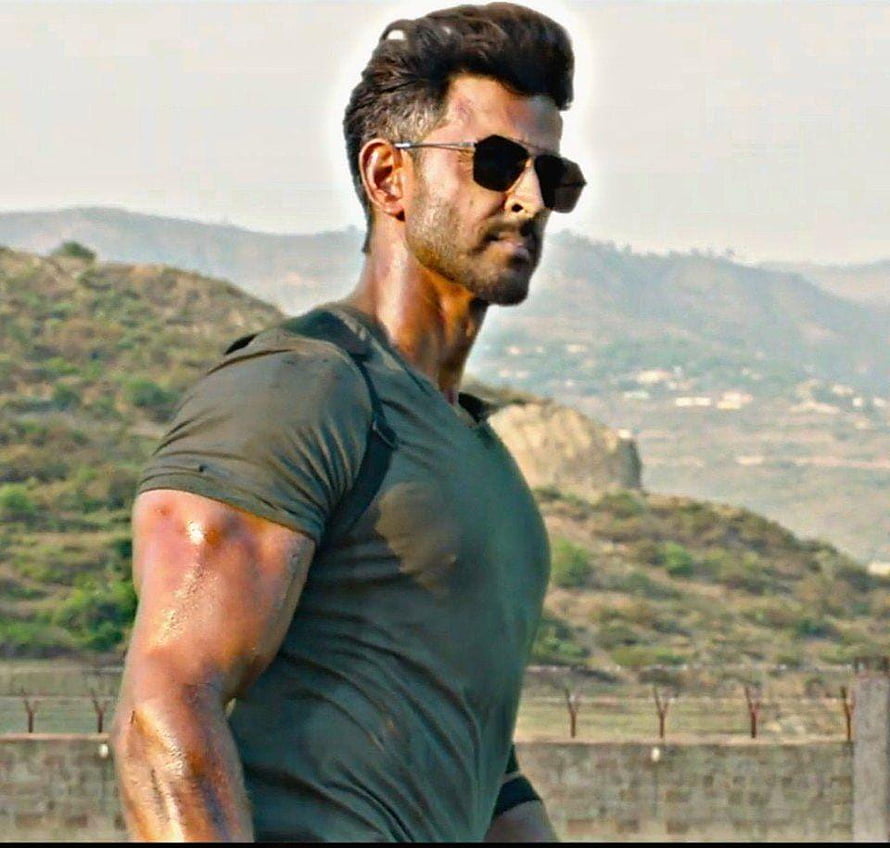 Hrithik Roshan in rugged military action pose with aviator sunglasses against a hilly landscape HD wallpaper
