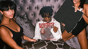 Juice WRLD in a white graphic tee with two women on a tufted bed HD wallpaper