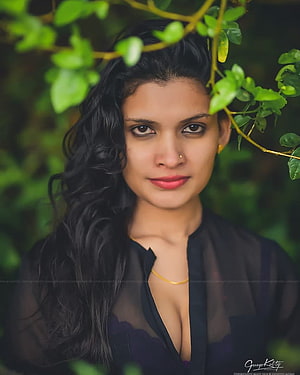 Portrait of a woman with long black wavy hair wearing a sheer black top and gold necklace amidst green foliage, Model Resmi R Nair New hoot, reshmi nair HD phone wallpaper