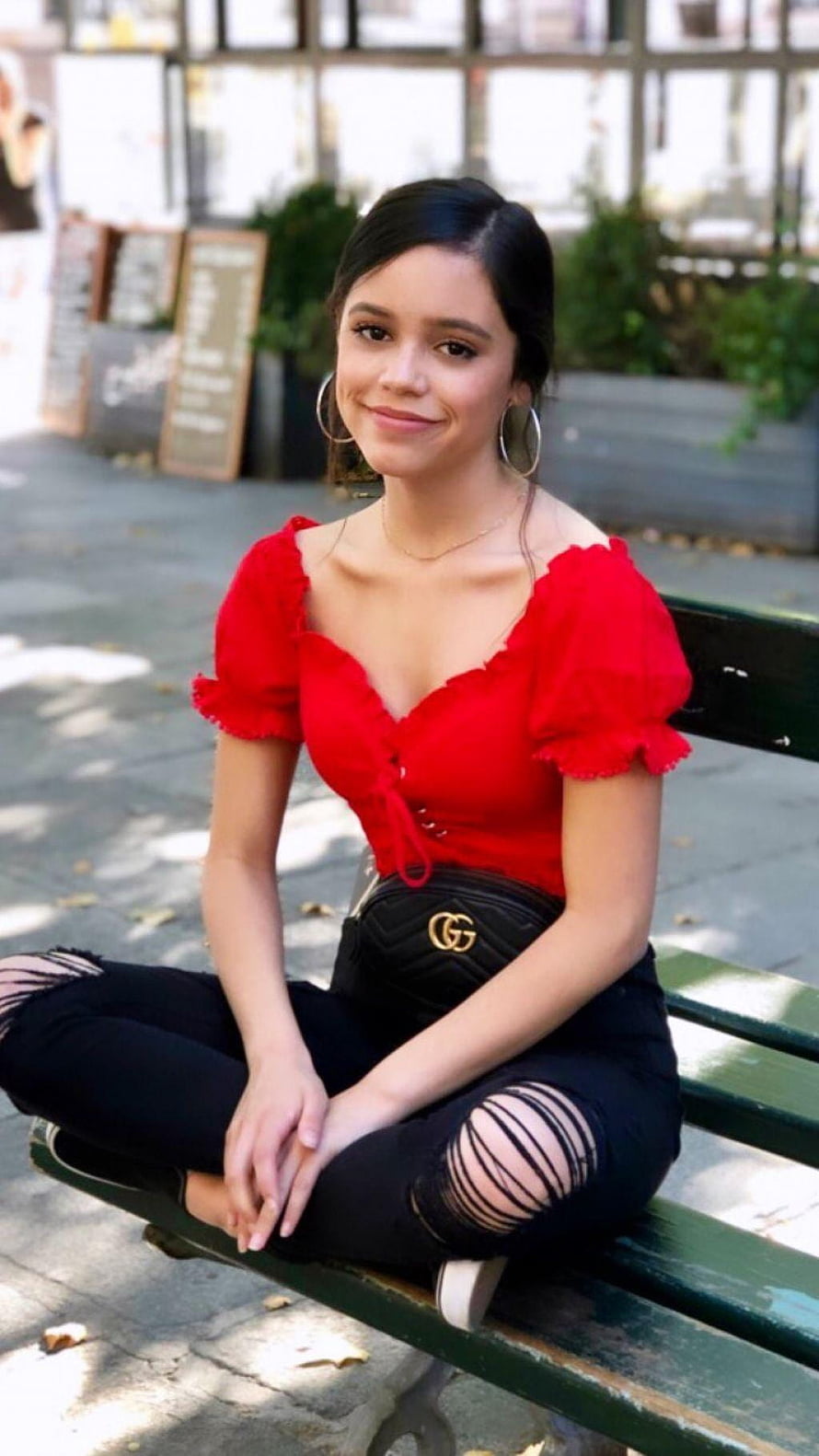 Jenna Ortega in a red ruffled top and black ripped jeans sitting on a park bench HD phone wallpaper