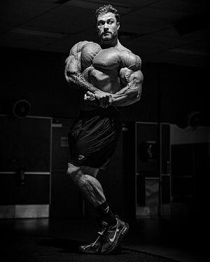 Black and white photo of a muscular bodybuilder posing in a gym with intense lighting HD phone wallpaper