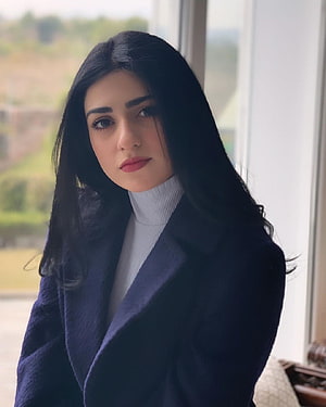 Sarah Khan in a navy coat and white turtleneck by a window HD phone wallpaper