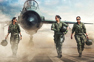 Three female fighter pilots in green flight suits walking confidently on a dusty runway with a military jet in the background HD wallpaper
