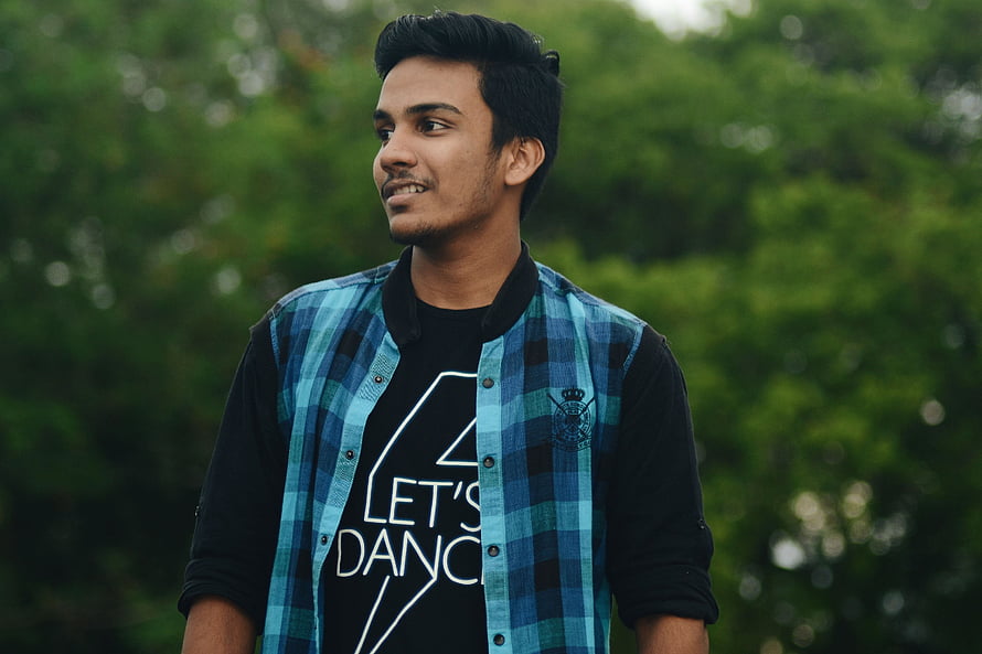 Young man in blue plaid shirt and black t-shirt standing outdoors with green foliage background, indian boy pic, #Indian Boy HD wallpaper