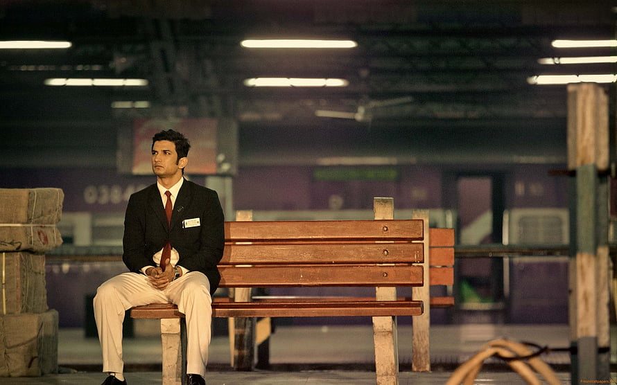 Sushant Singh Rajput as MS Dhoni in a formal suit sitting on a wooden bench at a train station platform HD wallpaper