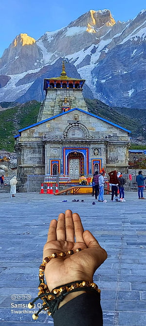 Hand holding prayer beads in front of Kedarnath Temple with snow-capped mountains HD phone wallpaper