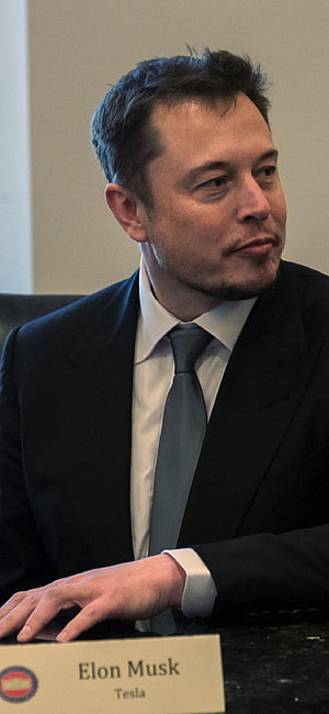 Elon Musk in a dark suit and grey tie seated at a formal meeting table with a nameplate HD phone wallpaper