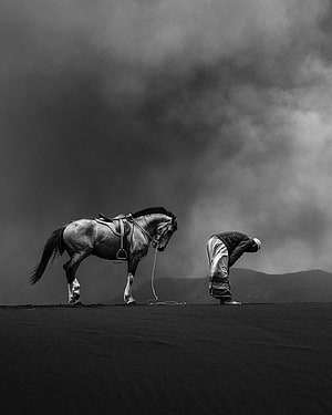 Samir Latif Black and White Photograph of a Man Praying Beside a Saddle Horse Under a Dramatic Sky, Samir Latif, horse and man praying HD phone wallpaper