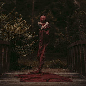 Eerie figure wrapped in flowing red fabric standing on a rustic wooden bridge in a dark forest HD phone wallpaper