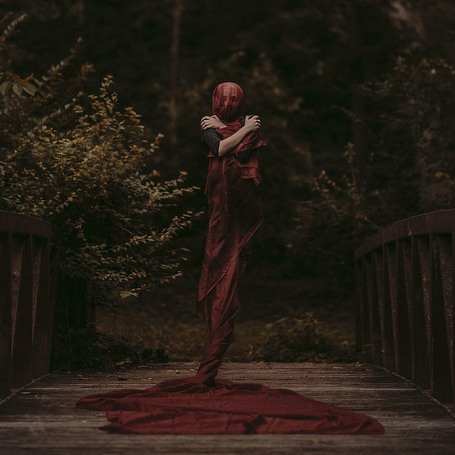 Eerie figure wrapped in flowing red fabric standing on a rustic wooden bridge in a dark forest HD phone wallpaper