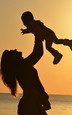Silhouette of a mother lifting her baby high against a golden sunset over the ocean HD phone wallpaper
