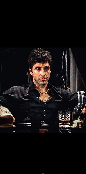 Tony Montana in a black shirt seated at a table with a whiskey glass and decanter in a dimly lit room HD phone wallpaper
