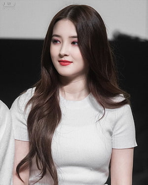 Nancy from Momoland in a white ribbed top with long wavy brown hair and soft studio lighting HD phone wallpaper
