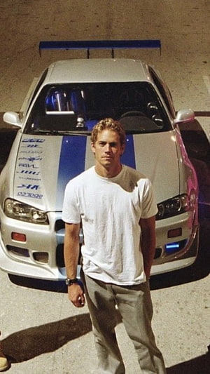 Paul Walker standing in front of the iconic silver Nissan Skyline GT-R R34 from the Fast and Furious movie franchise HD phone wallpaper