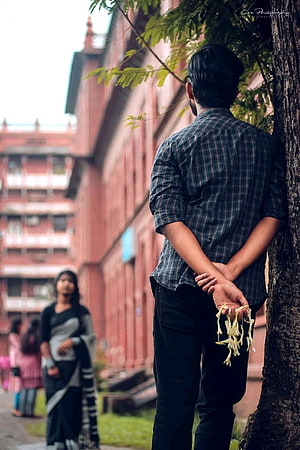 Man holding jasmine flowers behind back while leaning against tree with blurred woman in background, Pin on Bengali famous couple, bengali couples HD phone wallpaper