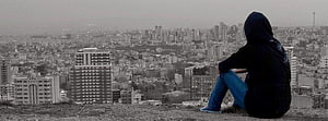 Hooded figure sitting on a hill overlooking a sprawling city skyline in muted tones HD wallpaper