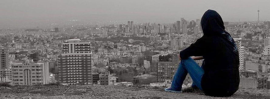 Hooded figure sitting on a hill overlooking a sprawling city skyline in muted tones HD wallpaper