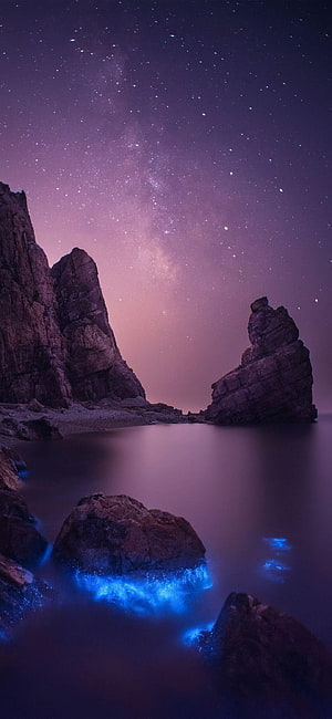Bioluminescent Waves Crashing on Rocky Shore Under Purple Milky Way Night Sky HD phone wallpaper