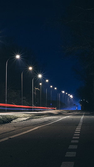 Nighttime street scene with glowing streetlights and red light trails on a dark road HD phone wallpaper