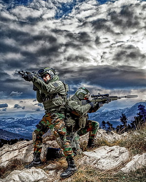 Two tactical soldiers aiming rifles on a rocky mountain ridge under a dramatic cloudy sky HD phone wallpaper