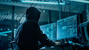 Anonymous Hacker in Hoodie Working on Multiple Computer Monitors in Dark Cyber Security Room HD wallpaper