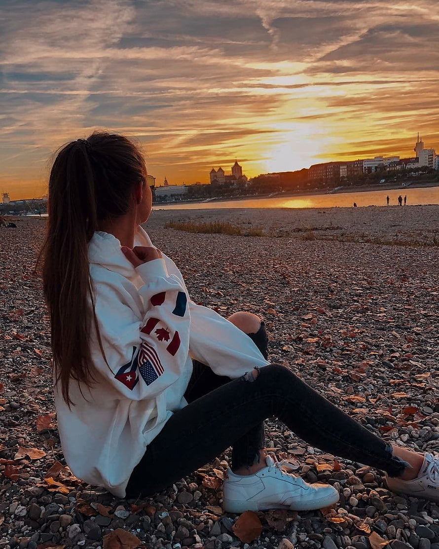 Girl in White Hoodie Sitting on Pebble Beach Watching Golden Sunset Over River City HD phone wallpaper