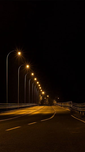 Empty asphalt road illuminated by glowing yellow streetlights at night HD phone wallpaper