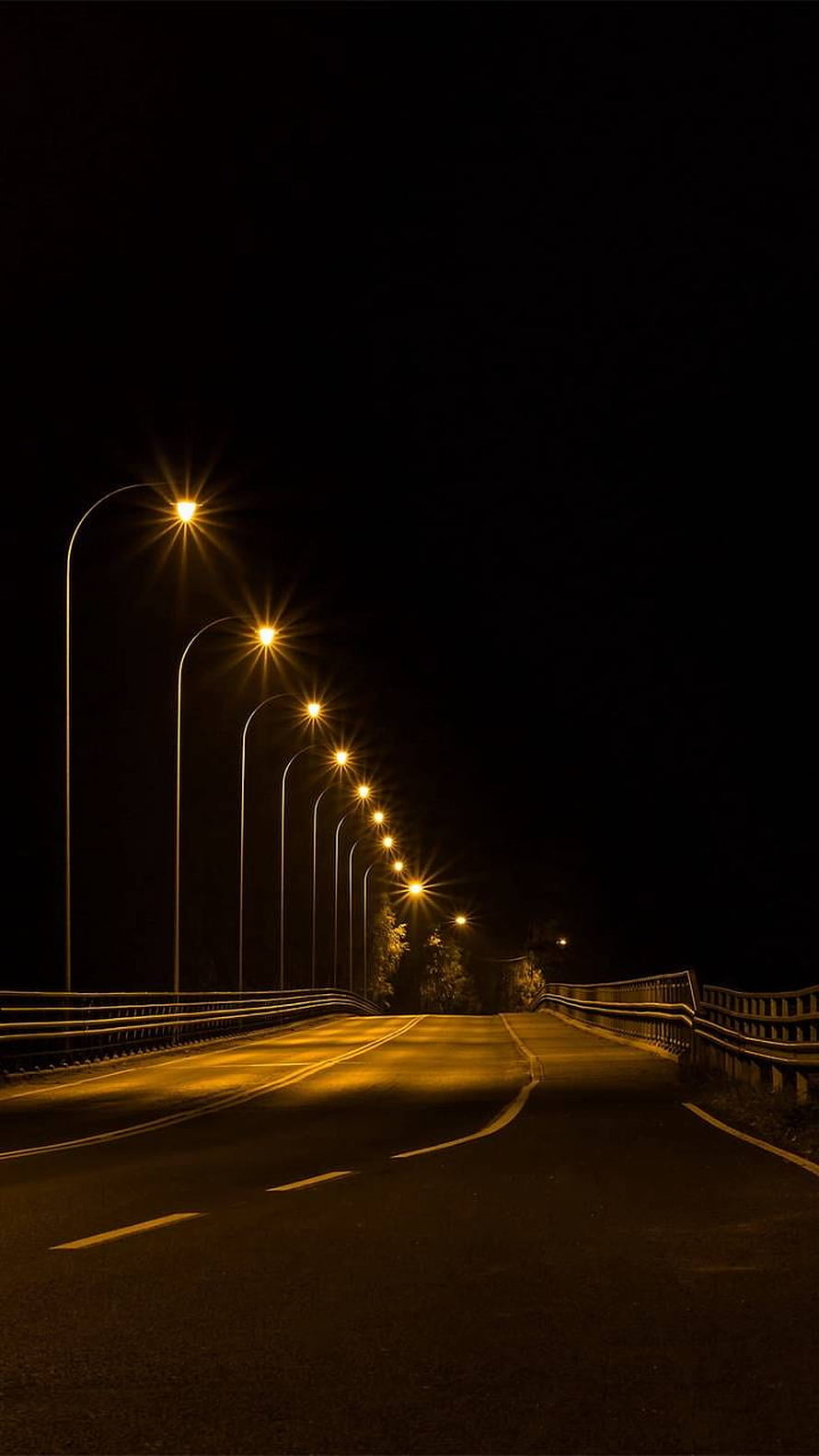 Empty asphalt road illuminated by glowing yellow streetlights at night HD phone wallpaper