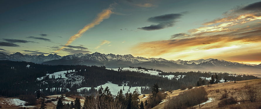 Panoramic Mountain Range at Golden Hour with Snowy Forests and Dramatic Sky HD wallpaper