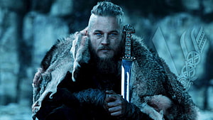 Ragnar Lothbrok in fur cloak holding sword in snowy forest from The Vikings series HD wallpaper