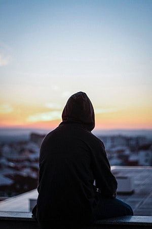 Silhouette of a person in a hoodie sitting on a rooftop overlooking a city at sunset HD phone wallpaper