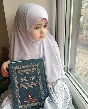 Baby girl in hijab holding a Quran translation book by a window HD phone wallpaper