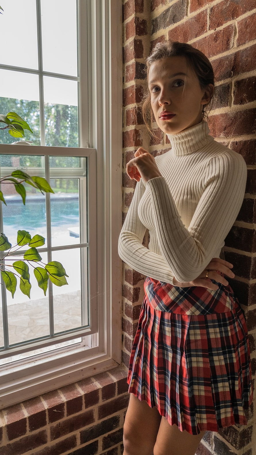 Millie Bobby Brown in a cream turtleneck and red plaid skirt by a brick window HD phone wallpaper