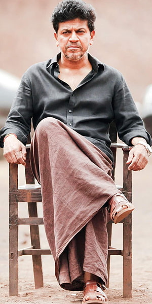 Shivarajkumar seated in traditional dhoti and black shirt on a rustic wooden chair HD phone wallpaper