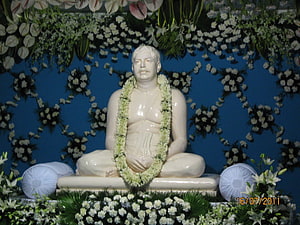 Sri Sri Thakur Anukulchandra seated statue adorned with a white flower garland against a blue floral backdrop HD wallpaper