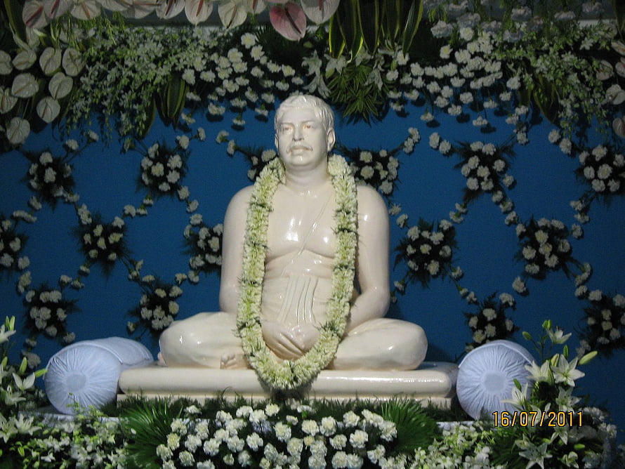 Sri Sri Thakur Anukulchandra seated statue adorned with a white flower garland against a blue floral backdrop HD wallpaper
